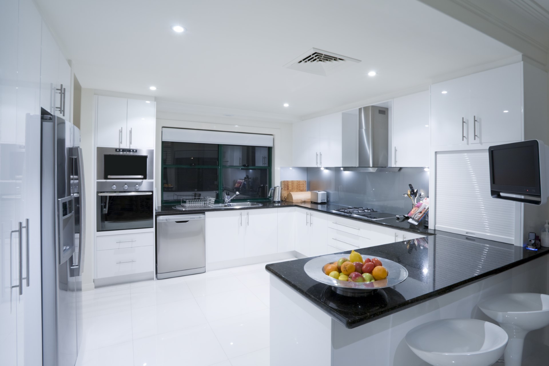 A modern kitchen with high gloss white cabinets, glossy black marble counter tops, glossy white tile floors, and stainless steel appliances. There is a bowl of fruit on the counter.